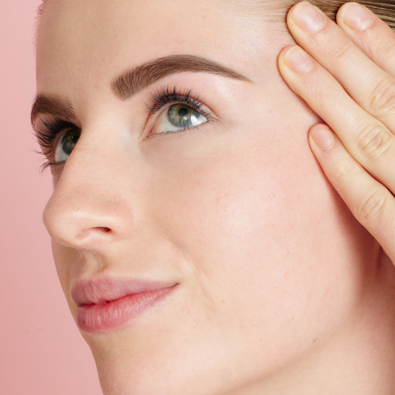 Close-up portrait of a woman with blonde hair and natural makeup, highlighting a neatly shaped and filled eyebrow, with her hand gently touching her temple against a soft pink background.