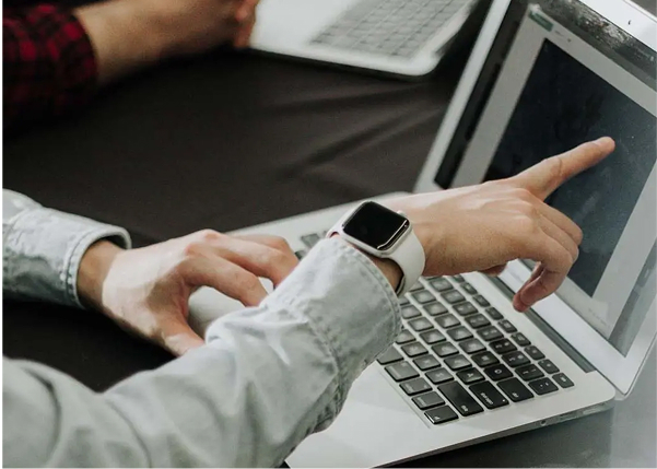 Close-up of a person's hands at a laptop, with one hand wearing a smartwatch and pointing at the screen, and the other hand resting on the keyboard.