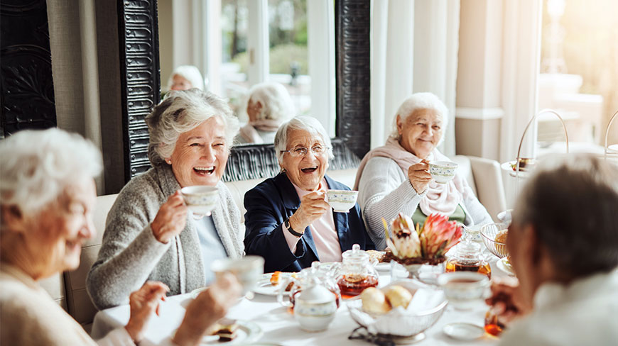 A group of smiling senior women enjoy afternoon tea together, laughing around a table in a sunlit room.