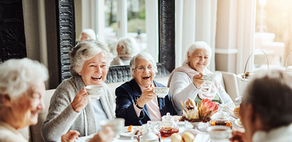 A group of smiling senior women enjoy afternoon tea together, laughing around a table in a sunlit room.