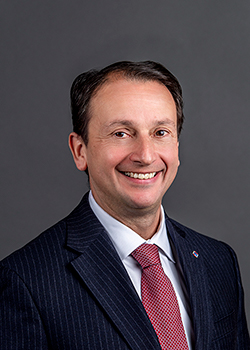 A portrait of Anthony Minopoli wearing a dark suit, white shirt, and red tie.