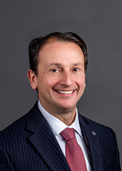 A portrait of Anthony Minopoli wearing a dark suit, white shirt, and red tie.