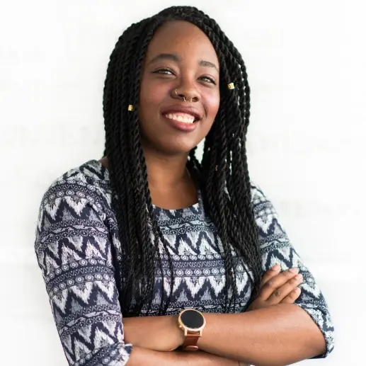 A smiling woman with long, braided hair and gold cuffs in her hair, wearing a patterned blue and white shirt and a smartwatch, with her arms crossed.