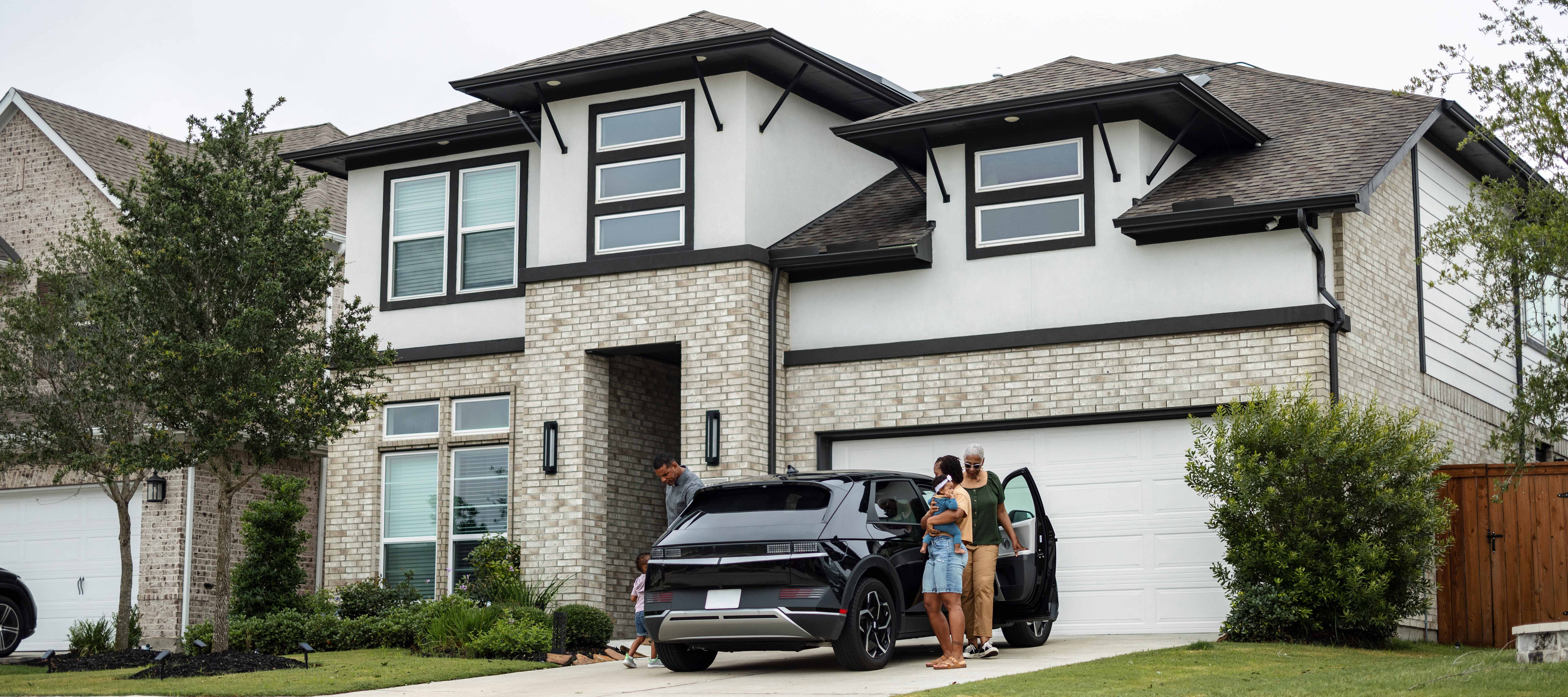 A Black multigenerational family gets out of a black SUV in the driveway of a nice white and black two story house