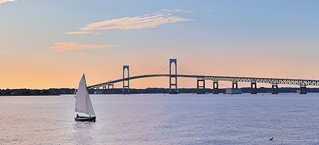 A sailboat sails in front of a bridge at sunset