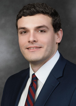 Headshot of Brendan Six, who smiles while wearing a dark blue suit and a red and blue striped tie