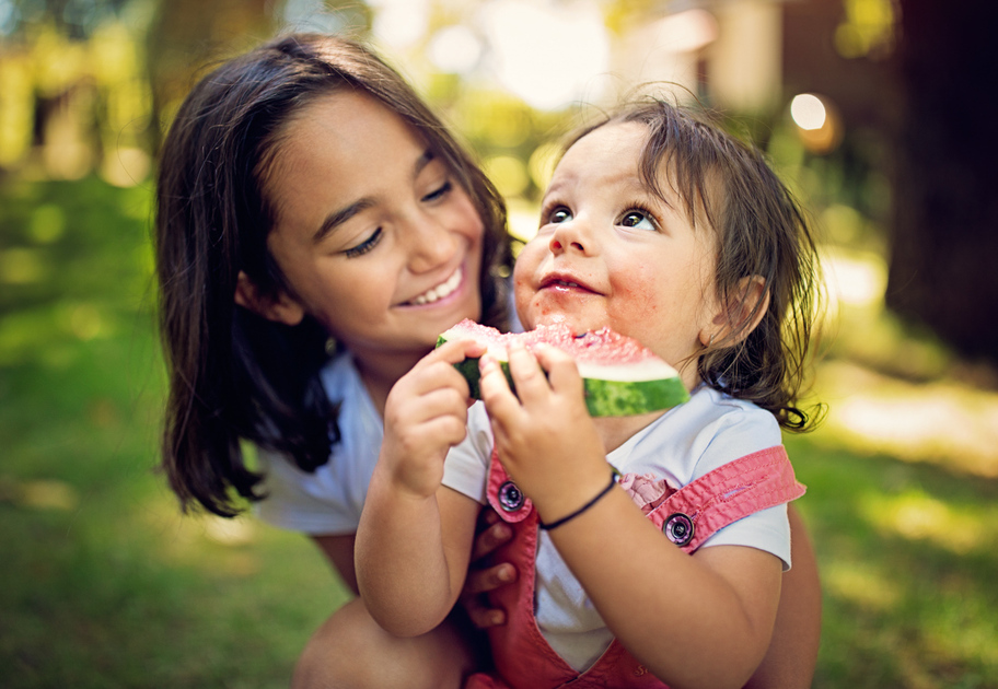 Two young girls are outside hugging. The older girl hugs her younger sister as she eats a watermelon.