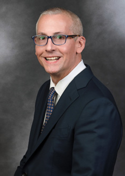 Headshot of Chris Kuen, who smiles while wearing a black suit and a blue tie