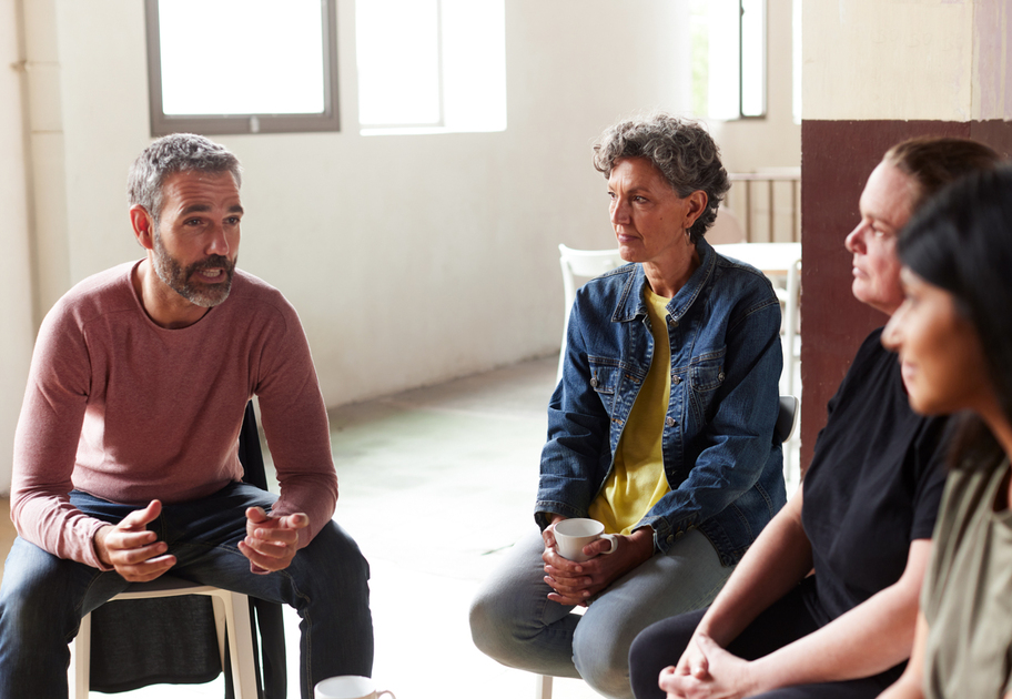 A man speaks while three women listen attentively in a bright room. All are seated in a circle, suggesting a group discussion or support meeting. Two women hold mugs, and natural light comes through the windows.