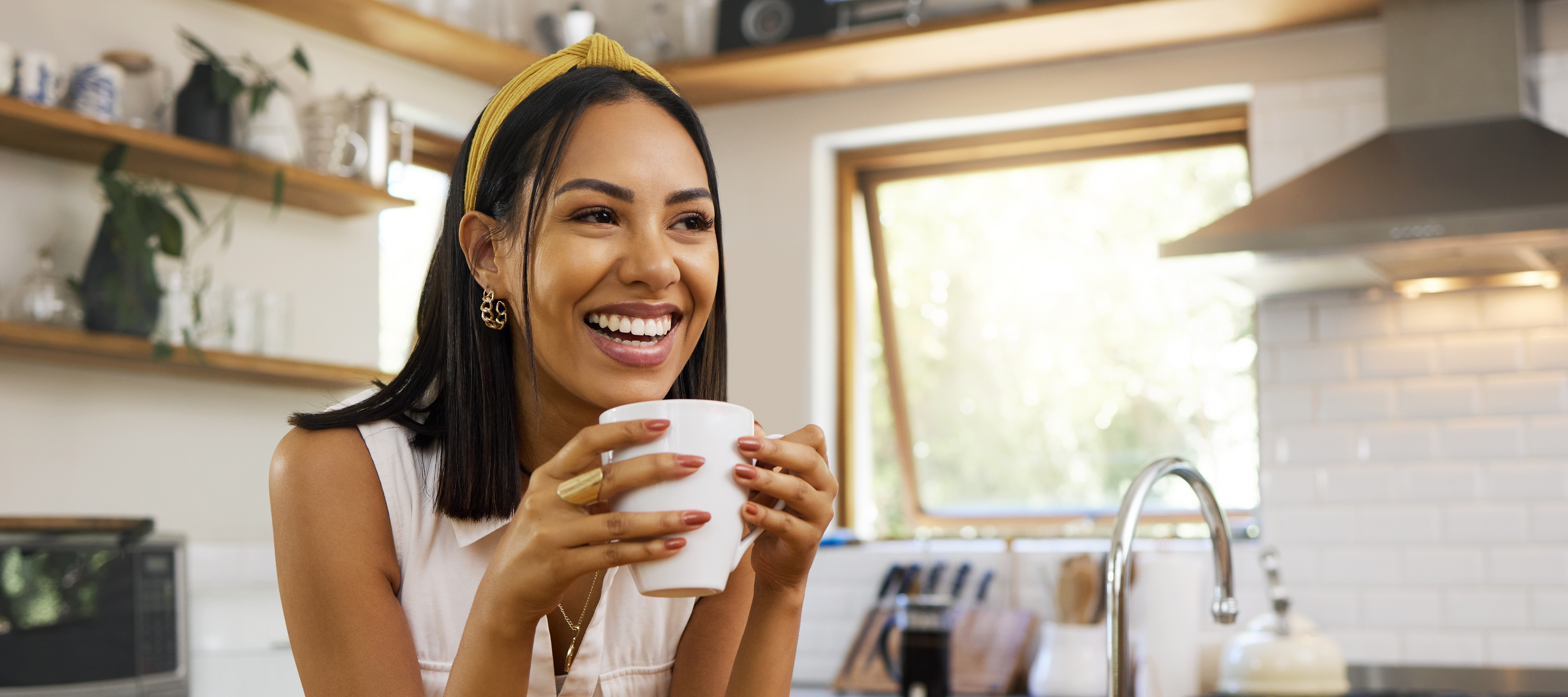A Latina woman smiles over her coffee cup while she sits at her kitchen table