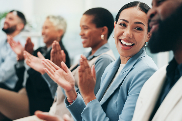 A diverse group of business professionals sitting in a row, smiling and applauding at an event. A woman in a light blue jacket in the foreground is looking forward and clapping enthusiastically.