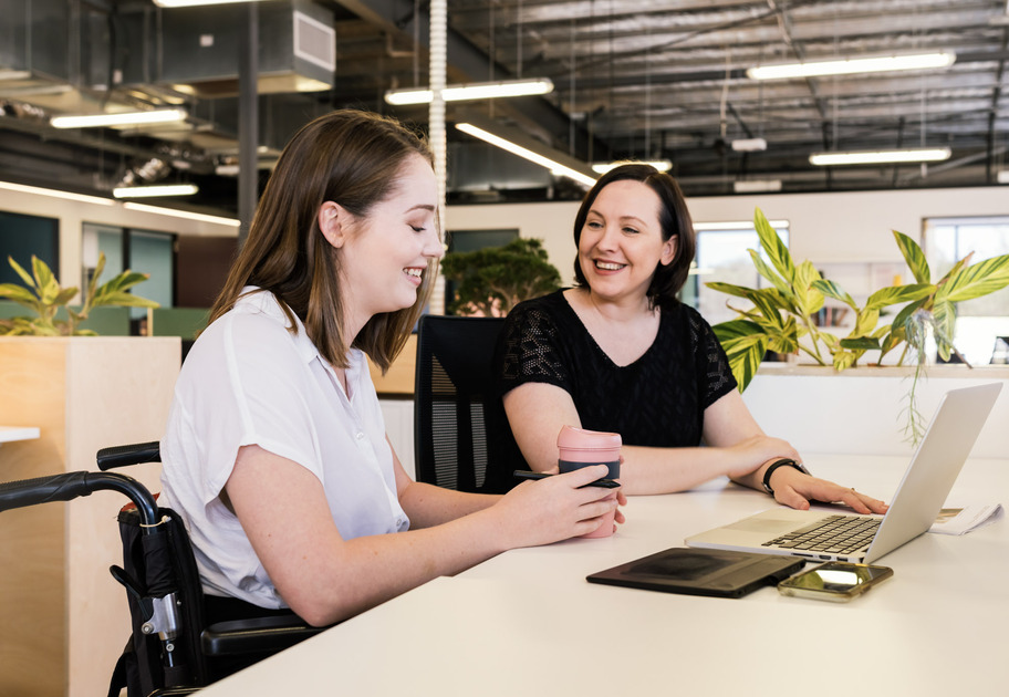 Two women in an office setting; the younger woman, who is in a wheelchair, is smiling as she looks down at a computer, possibly engaged in training. The older woman smiles in encouragement at the younger woman.