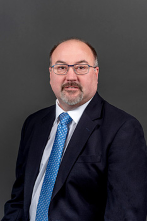 Headshot of Douglas Riley, who smiles while wearing a black suit and a blue tie