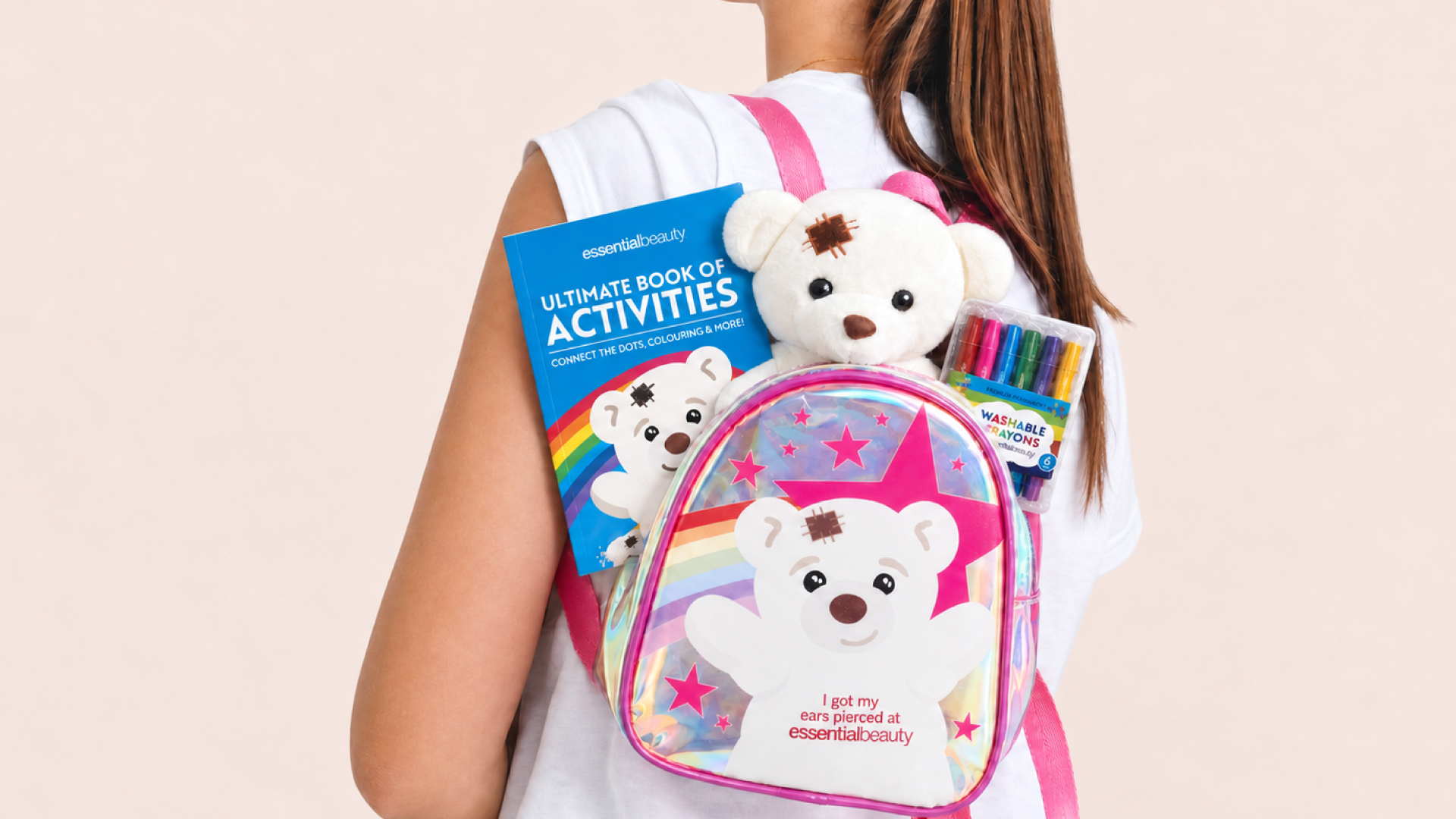 Person holding an Essential Beauty children’s ear-piercing backpack filled with a teddy bear, colouring activity book and washable markers.