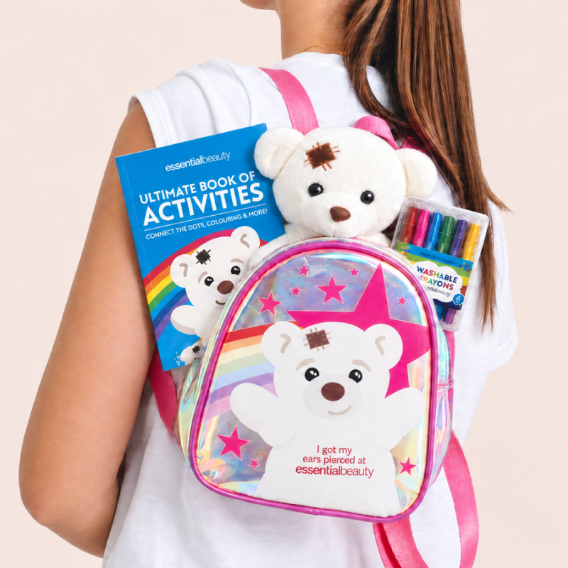 Person holding an Essential Beauty children’s ear-piercing backpack filled with a teddy bear, colouring activity book and washable markers.