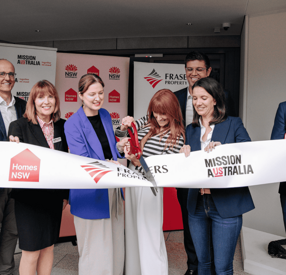 Seven individuals participate in a ribbon-cutting ceremony for a housing project, with banners and a ribbon displaying logos for NSW Government, Homes NSW, Frasers Property, and Mission Australia.