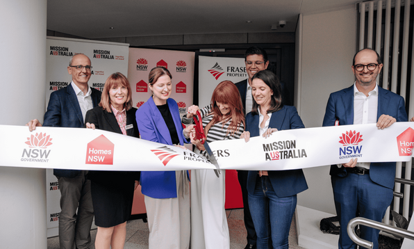 Seven individuals participate in a ribbon-cutting ceremony for a housing project, with banners and a ribbon displaying logos for NSW Government, Homes NSW, Frasers Property, and Mission Australia.