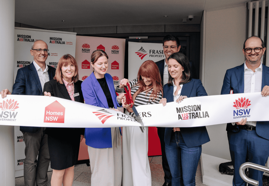 Seven individuals participate in a ribbon-cutting ceremony for a housing project, with banners and a ribbon displaying logos for NSW Government, Homes NSW, Frasers Property, and Mission Australia.