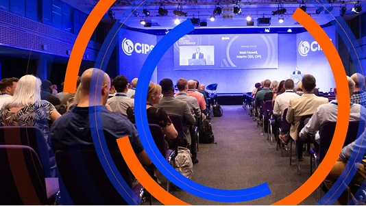 An audience is seated in rows facing a stage at a conference. A speaker is presenting on the stage, and the screen behind him displays his name: Matt Howell, Interim CEO, CIPS. The image is partially overlaid with abstract blue and orange circular lines.