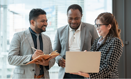 Three business professionals, two men and one woman, are standing and collaborating in a brightly lit modern office space. The man on the left is smiling and writing in a notebook, while the woman on the right holds a laptop and points at the screen. The man in the middle looks over her shoulder.