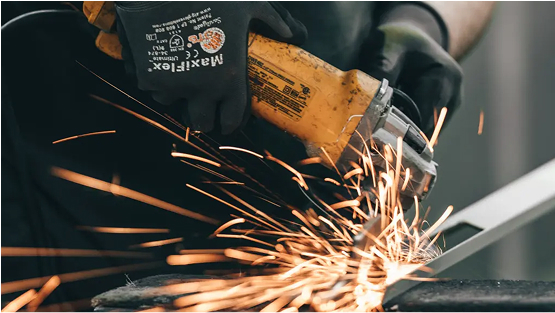 A close-up shot of a person wearing black protective gloves operating an angle grinder, creating a shower of bright orange sparks as they cut a piece of metal.