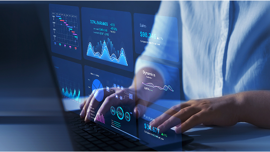 Close-up of a person's hands typing on a laptop keyboard, with futuristic blue holographic screens displaying various business data and financial charts floating above the keyboard.