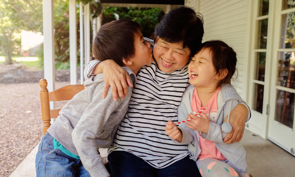 An older asian lady embraces a young boy and young girl. They are laughing and smiling as the young boy kisses the lady on her cheek. 