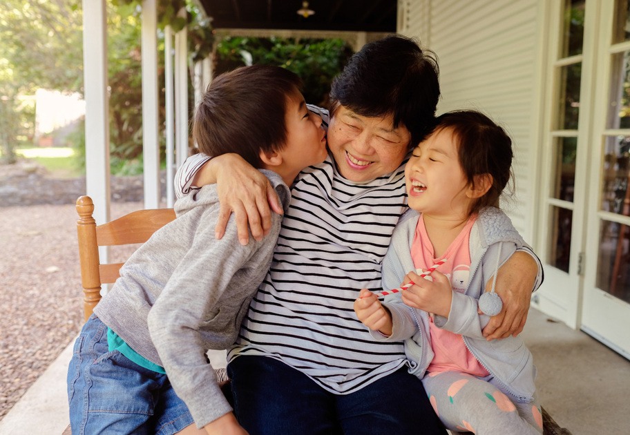 An older asian lady embraces a young boy and young girl. They are laughing and smiling as the young boy kisses the lady on her cheek. 