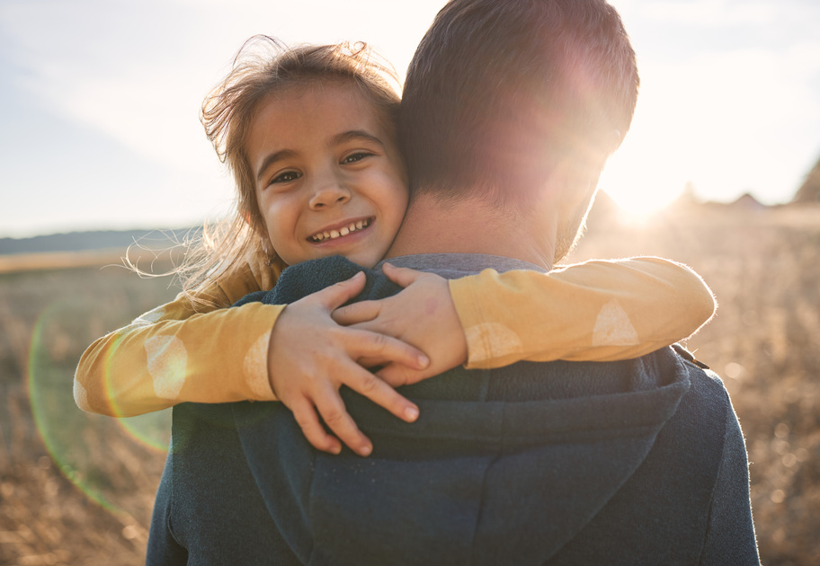 Daughter hugs her father in a big hug and smiles at the camera. Her father is facing a field that is bathed in sunlight. 