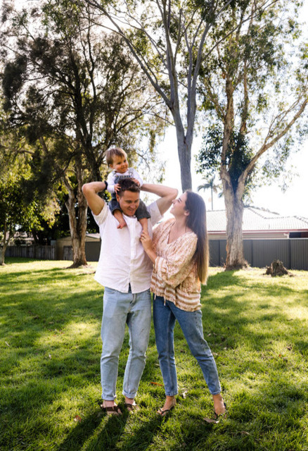 A man stands in a grassy park holding a child on his shoulders, while a woman stands beside them, smiling and touching the child. Trees and houses are visible in the background.