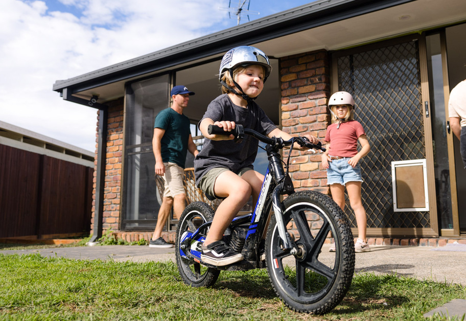 A young child wearing a helmet rides a bicycle on a lawn outside a house, while two adults and another child stand nearby.