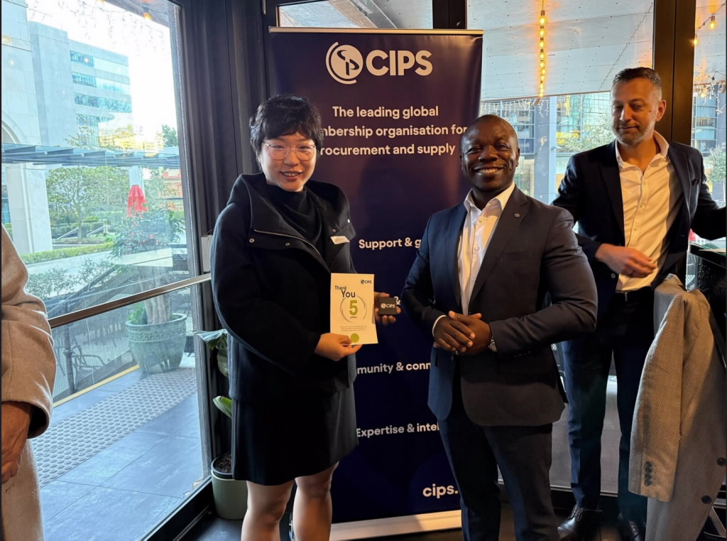 A woman in a black jacket is smiling and holding a CIPS five-year volunteer award certificate and card. She is standing next to a man in a dark suit, also smiling. A CIPS banner is centred behind them, and another man in a white shirt and dark jacket is visible to the right.