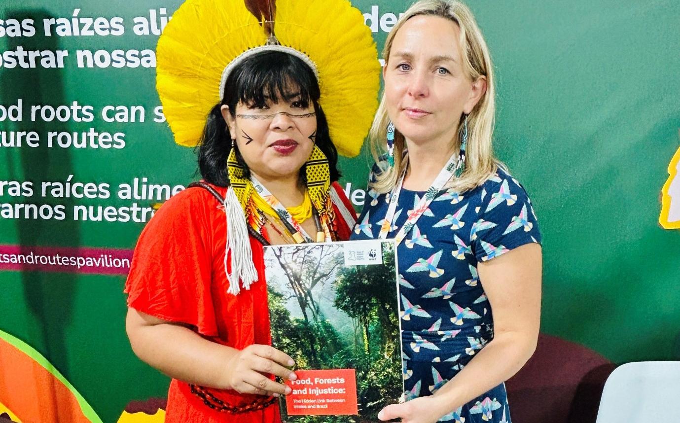 Two women stand together holding a book titled 'Food, Forests and Injustice' in front of a green display with multilingual text.

