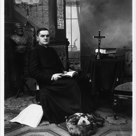 Father Michael J. McGivney seated in studio rectory setting, dressed in a long cassock, seated in high-backed upholstered chair, opened book in lap.