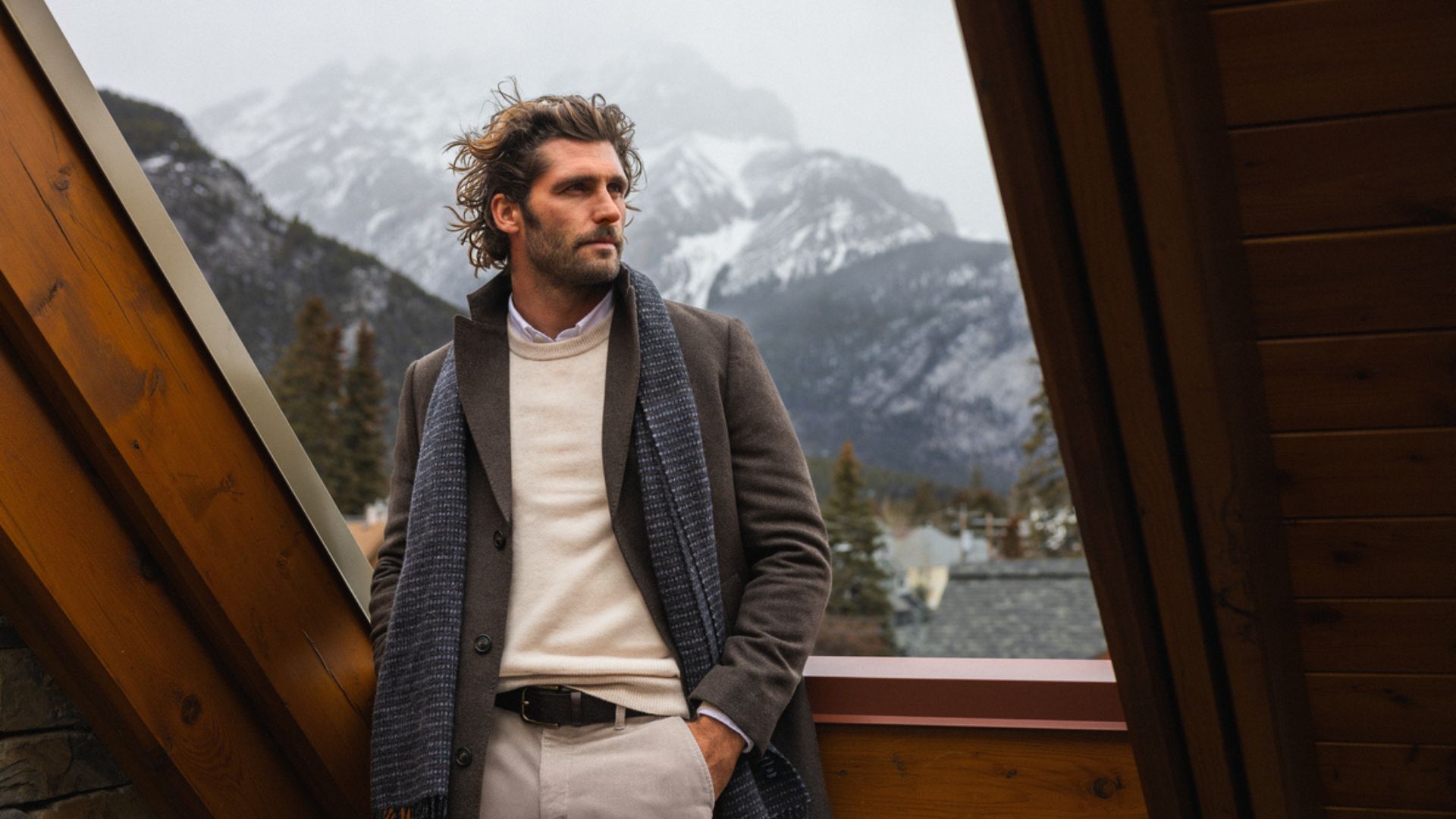 Person wearing a brown overcoat, beige knit jumper, trousers and a dark scarf, standing on a timber balcony with a mountainous, snow-covered landscape in the background.