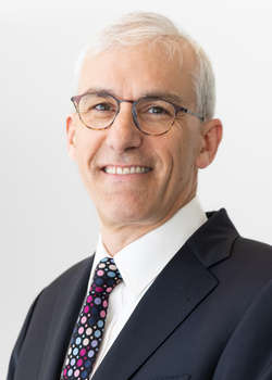 A corporate headshot style portrait of Grattan Brown, smiling wearing a dark suit, white shirt, and pink and teal polka dotted tie on a light grey background.