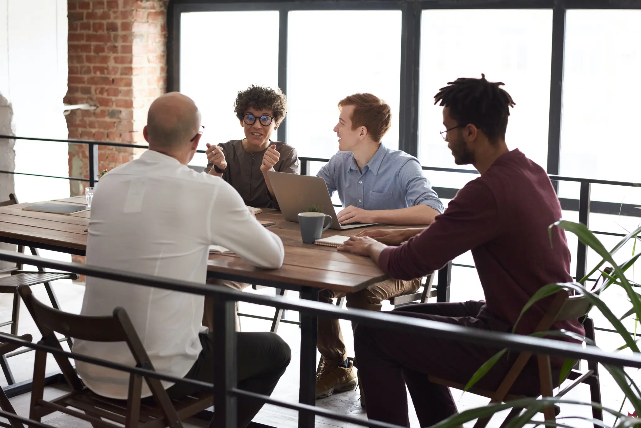 Four men are seated around a long, rustic wooden table in a brightly lit modern office setting. They appear to be having a casual business meeting or discussion. The men on the right side of the table are facing the men on the left. The man in the foreground on the left has his back to the viewer, wearing a white long-sleeve shirt. The other three men are engaged in conversation; one is wearing glasses and gesturing while speaking, another is smiling in a blue shirt next to an open laptop, and the third is listening in a deep red long-ssleeve shirt. The office features exposed brick walls and large, floor-to-ceiling windows that provide strong backlighting.