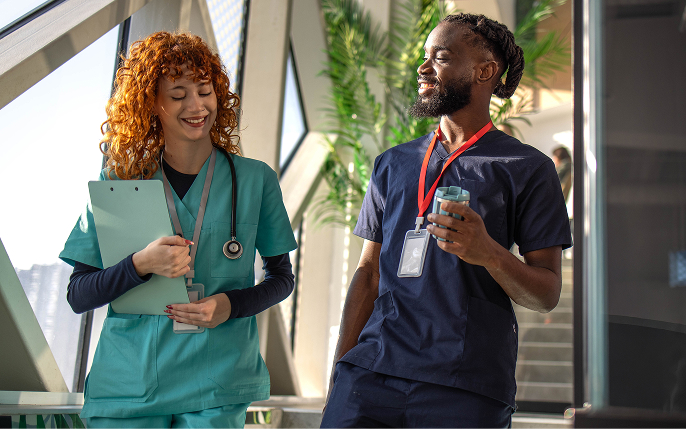 Two healthcare workers in medical scrubs walk down a modern hallway with large glass windows and indoor plants, smiling and talking. The worker on the left wears teal scrubs and carries a clipboard with a stethoscope around her neck; the worker on the right wears navy scrubs and holds a travel mug.