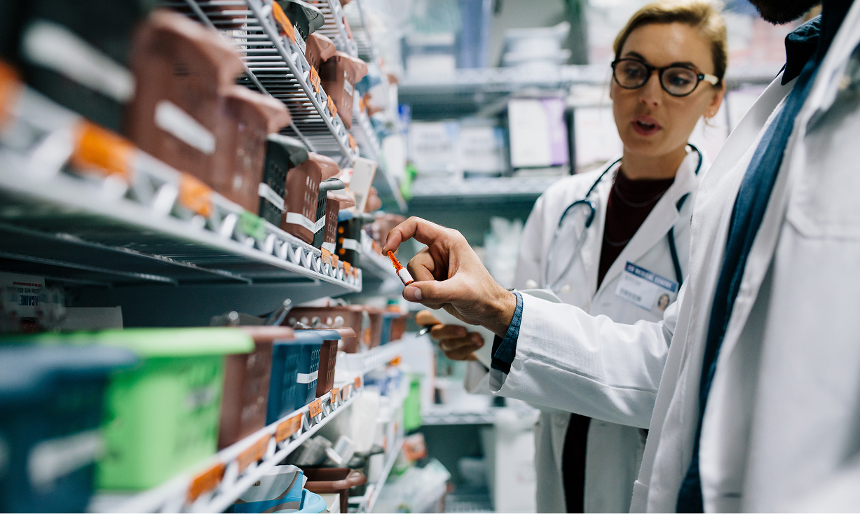 A close-up shot of two healthcare professionals in white lab coats standing next to organized medical storage shelves. One professional's hand is in focus, carefully holding up a small orange medication ampoule for inspection.