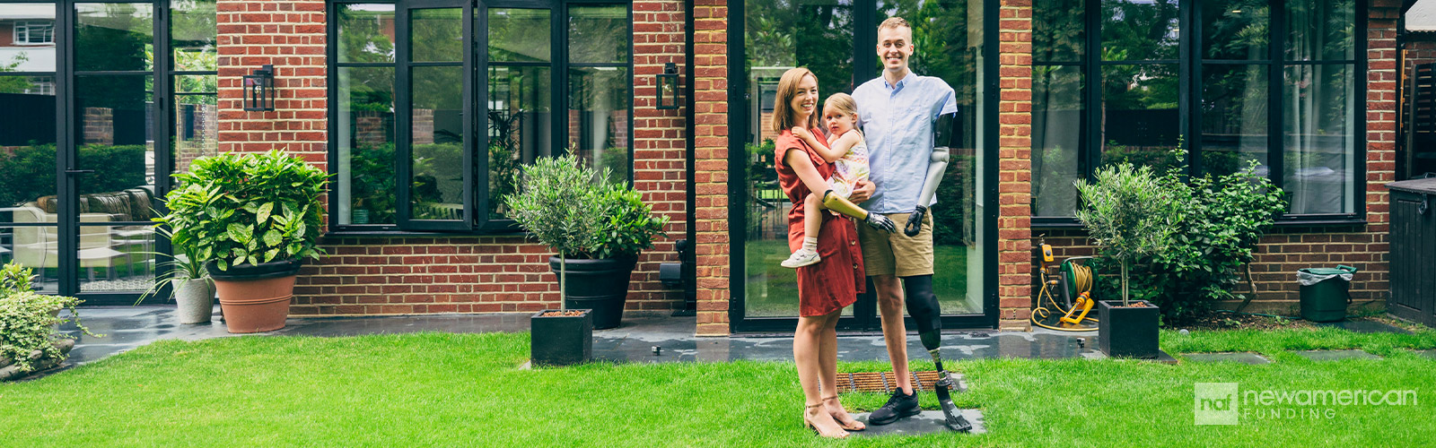 A disabled couple stands smiling with their child in front of their new home