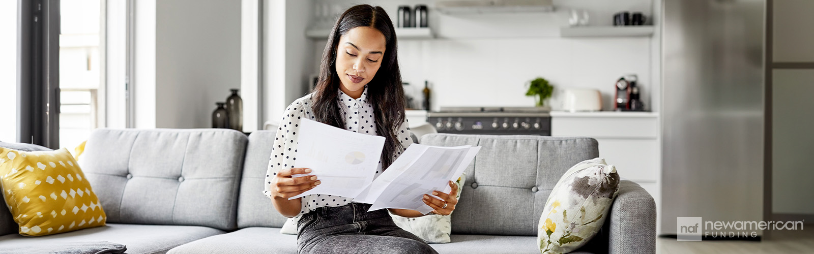 A Black woman sits on a couch in a home holding and comparing two sheets of paper with information on them