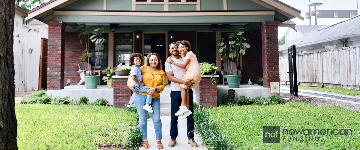 A smiling Latina woman stands with a smiling Black man and two smiling children in front of a nice home