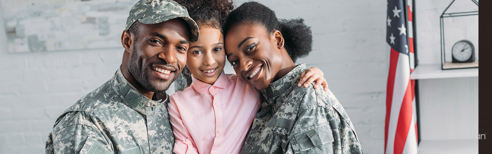 A Black man and Black woman dressed in military uniforms stand smiling on either side of a Black girl with an American flag behind them