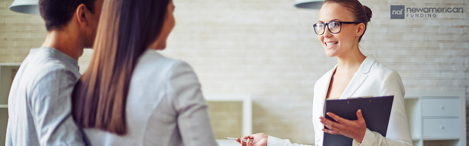 A white woman in a blazer is smiling and holding a clipboard while she talks to a man and woman inside of a house