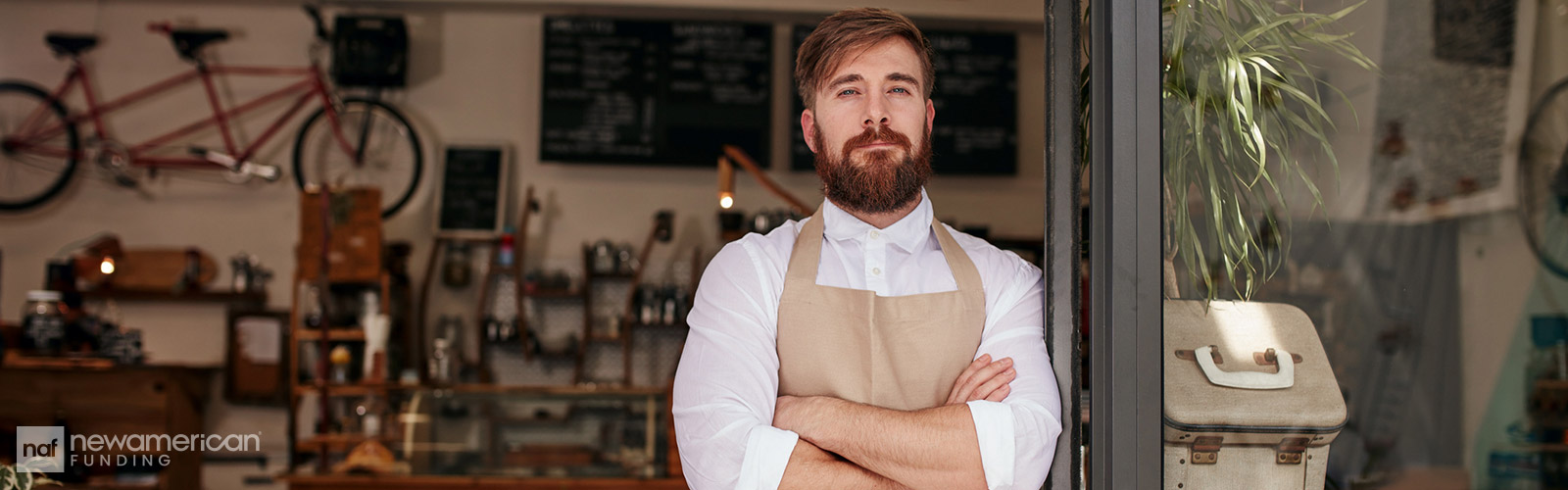 A white man stands in front of a shop wearing a white button down and a brown apron