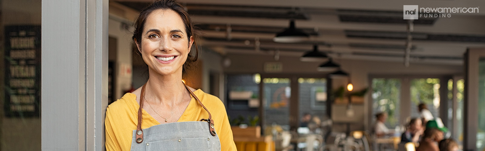 A Hispanic woman stands in front of a busy restaurant wearing an apron
