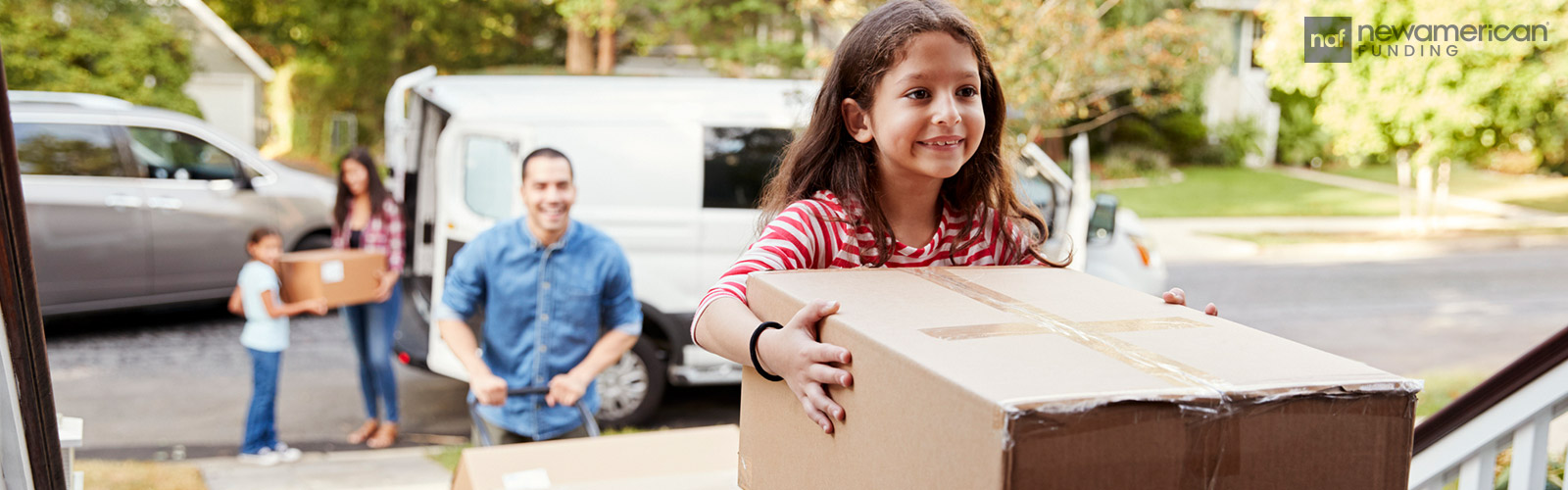 A little smiling Hispanic girl carries a moving box into a home while her parents walk behind her