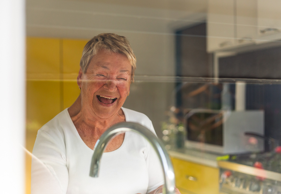 An elderly woman with short gray hair smiles and laughs while standing in a bright kitchen, seen through a window near a shiny kitchen faucet.