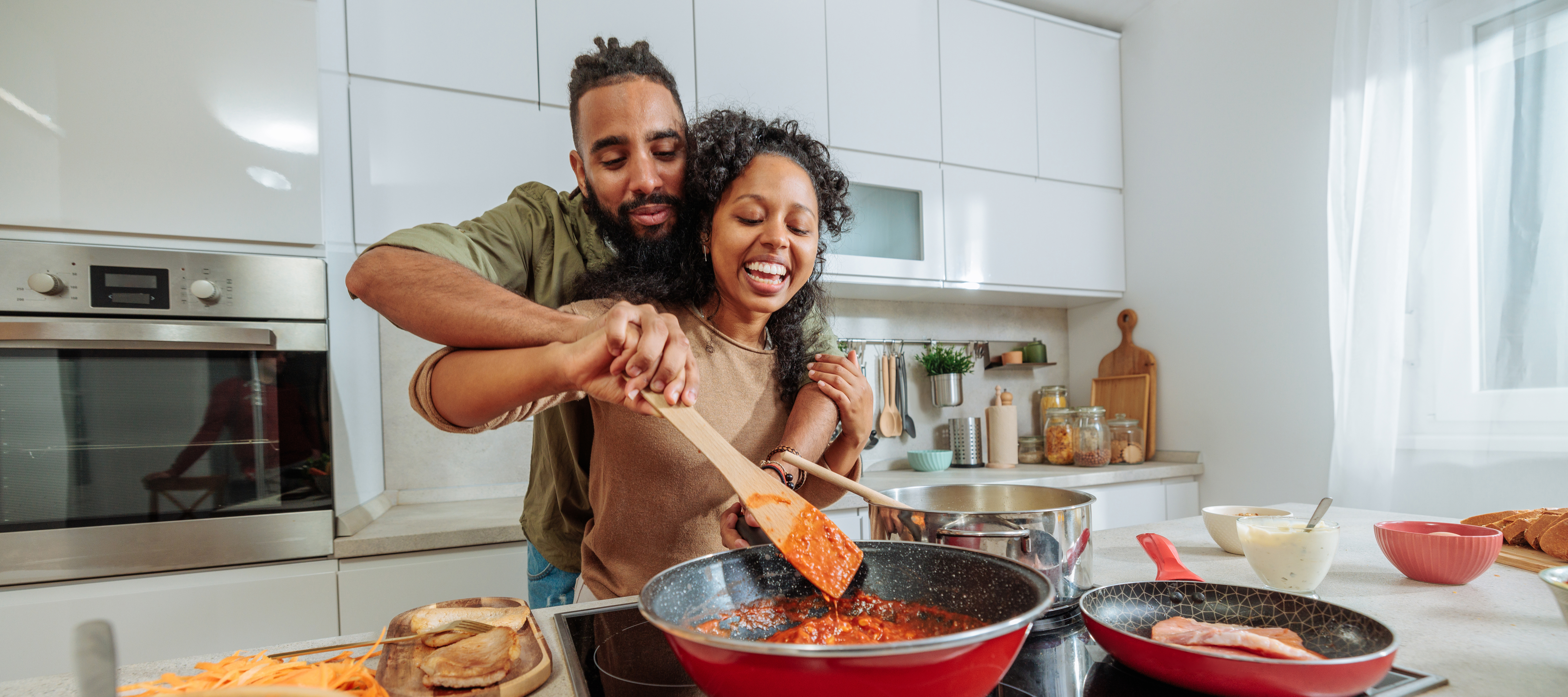 A Black woman and man laugh together while they cook in their kitchen