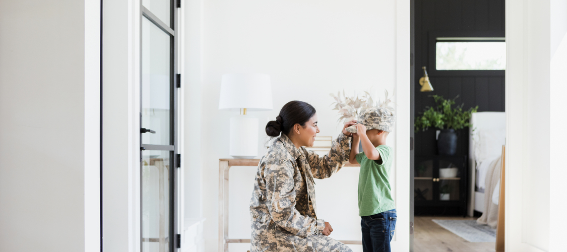 A woman in a military uniform at a home putting her hat on a small boy.